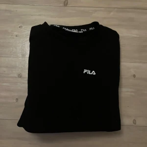 Svart sweatshirt från FILA - Svart sweatshirt från FILA med vit broderad logga på bröstet. Tröjan har en klassisk rund hals och är tillverkad i mjuk bomull och polyester, vilket gör den skön att bära. Perfekt för en chill och sportig stil.