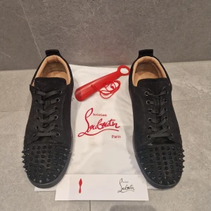 Christian Louboutin svarta sneakers med nitar - Svarta sneakers från Christian Louboutin i mocka med ikoniska röda sulor. Skorna har rund tå, snörning och är dekorerade med svarta nitar på tån för en edgy look. Insidan är i beige skinn och sulan har Louboutins signatur. Perfekta för dig som vill sticka ut.