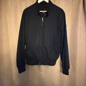 Marinblå vind från J.Lindeberg - Stilren svart bomberjacka från J.Lindeberg med klassisk passform. Jackan har dragkedja framtill, ribbade muddar och två fickor med knappstängning. Snygg logotyp på ärmen och mjukt, lätt material som passar perfekt till vardagslooken.