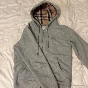 Grå hoodie från Burberry med dragkedja - Snygg grå hoodie från Burberry med klassiskt rutigt foder i huvan och broderad logga på bröstet. Tröjan har dragkedja framtill, känguruficka och snörning i huvan. Tillverkad i mjuk bomull med lite stretch för extra komfort.
