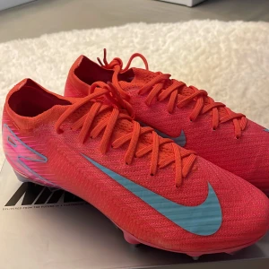 Nike Mercurial fotbollsskor röd/blå - Säljer ett par Nike Mercurial fotbollsskor i en riktigt snygg röd färg med blå detaljer och logga. Skorna har snörning och är tillverkade i lätt syntetmaterial för bästa känsla på planen. Rund tå och platt sula med dobbar för gräs. Med följer matchande röd/blå fotbolls påse för skorna och extra metall dobbar med ett slags verkty.Original pris 3499kr