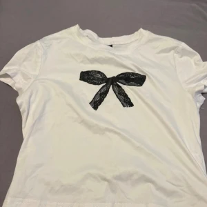 Vit t-shirt med svart rosett från SHEIN - Snygg vit t-shirt från SHEIN i storlek M med en stor svart rosett framtill. T-shirten har rund hals och korta ärmar, perfekt för en chill och trendig look. Materialet känns mjukt och lätt mot huden. Är en magtröja 