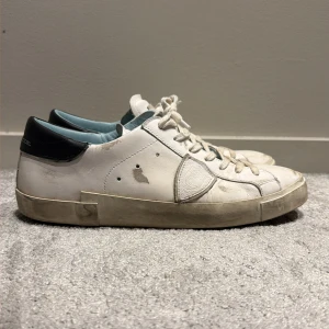 Philippe Model vita sneakers i skinn - Snygga vita sneakers från Philippe Model med svart häl och ljusblå insida. Skorna har rund tå, snörning och platt sula. Tillverkade i Italien i mjukt skinn med diskret logodetalj på sidan. Perfekta för dig som gillar stilrena och klassiska sneakers. Mitt pris 1200 nypris 3800