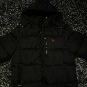 Svart pufferjacka Polo Ralph Lauren - Svart dunjacka från Polo Ralph Lauren med röd broderad logga på bröstet. Jackan har huva, dragkedja framtill och två sidofickor med dragkedja. Storlek M passar även L. Perfekt för kalla dagar. Kvitto finns. Bor i Göteborg.