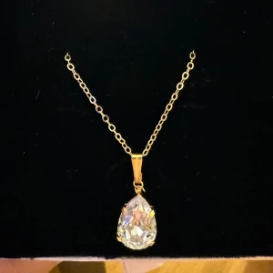 Guldhalsband med droppformad kristall - Snyggt halsband med tunn guldfärgad kedja 50cm och en stor, droppformad Swarovski kristall storlek 10mm som hänge. Kedjan har ovala länkar och hänget sitter i ett stilrent fäste. Perfekt för dig som gillar bling och vill sticka ut med något glittrigt och elegant.