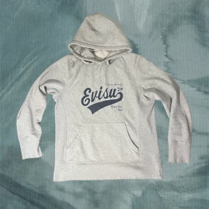 Grå hoodie från Evisu med tryck - Snygg ljusgrå hoodie från Evisu med stort mörkblått logotryck på bröstet och japanska tecken. Klassisk känguruficka framtill och huva. Mjuk bomullskvalitet som är skön att ha på sig. Perfekt för en chill och avslappnad stil. Snygg vintage slitage som ackumulerats med tiden. En av mina favoriter och självklart äkta. Ha det gött! // Gidlöfs