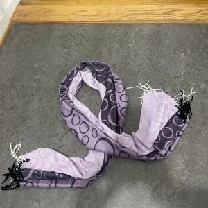 Ljuslila halsduk med cirkelmönster - Snygg ljuslila halsduk med svarta och vita cirklar i olika storlekar. Halsduken har fransar i svart och vitt i ändarna och är tillverkad i ett mjukt, tunt material som känns skönt mot huden. Perfekt accessoar för att lyfta din outfit.