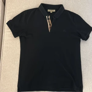 Svart Burberry piké - Snygg svart t-shirt från Burberry med klassisk krage och korta ärmar. Har den ikoniska Burberry-rutiga detaljen vid knäppningen fram. Materialet känns mjukt och skönt, perfekt för en clean och stilren look.