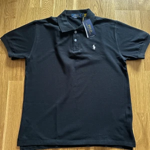 Svart pikétröja från Polo Ralph Lauren - Svart pikétröja från Polo Ralph Lauren med klassisk krage och två knappar framtill. Tröjan har korta ärmar och den ikoniska broderade loggan i vitt på bröstet. Tillverkad i mjuk bomull som känns skön mot huden. Perfekt för dig som gillar stilren och tidlös design.