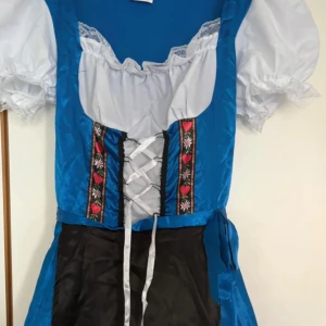 Blå October fest klänning - Snygg blå Octoberfest klänning från Boland med vit puffärm, svart förkläde och dekorativa band med röda hjärtan och blommor. Snörning framtill och vita spetsdetaljer vid hals och ärmslut. Kjolen har vita ränder och är tillverkad i glansig polyester. Köpt här på plick men aldrig använd av mig.