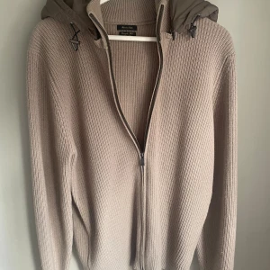 Beige stickad hoodie från Massimo Dutti - Säljer en beige stickad hoodie från Massimo Dutti i extrafin ull. Tröjan har en ribbad struktur, dragkedja framtill och en quiltad huva med snörning. Perfekt för dig som gillar stilrena och mysiga plagg med snygga detaljer. Denna varan är mycket eftertraktad eftersom den inte sälj längre