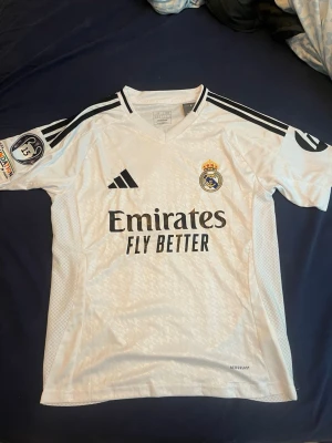 Real Madrid Mbappé matchtröja Adidas S - Säljer en vit Real Madrid matchtröja från Adidas med Mbappé och nummer 9 på ryggen. Tröjan har svarta detaljer, klubbmärke på bröstet och Champions League-märke på ärmen. Materialet är lätt polyester.