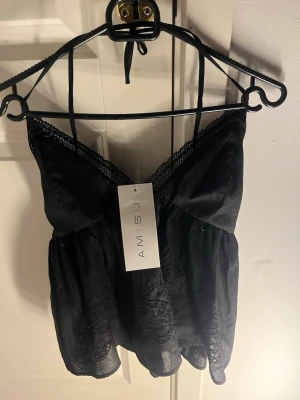 Svart broderad linnetopp från Amisu - Säljer en svart linnetopp från Amisu med tunna axelband som knyts i nacken. Toppen har spetskant vid v-ringningen och broderade detaljer nertill. Materialet är lätt och luftigt, perfekt för varma dagar. Snygg och feminin design med fina detaljer.