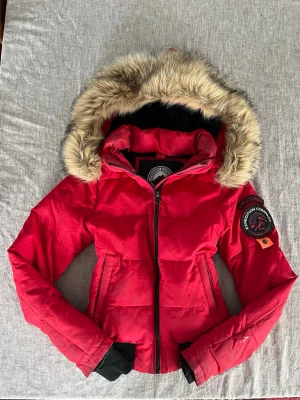 Röd pufferjacka från Superdry - Säljer en röd pufferjacka från Superdry med fluffig fuskpäls på huvan och coolt Expedition Company-märke på ärmen. Jackan har svarta ribbade muddar, dragkedja framtill och två fickor. Perfekt för kalla dagar och riktigt snygg streetstil.lite sliten (kolla bilder)