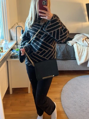 Svart axelväska med guldkedja - Snygg svart axelväska med stilren rektangulär form och guldfärgad kedja som axelrem. Väskan är gjord i skinnimitation och har en enkel, elegant design med ett metallspänne framtill. Perfekt storlek för mobil, plånbok och småsaker. Vet ej vart den är ifrån 