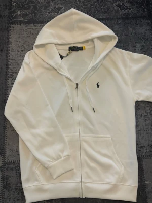Vit Ralph lauren zip up - Klassisk och tidlös design med den ikoniska broderade Ralph Lauren-loggan på bröstet. Hoodien är tillverkad i mjukt och bekvämt material med huva och justerbara snören samt känguruficka framtill. Passar perfekt till både vardag och mer uppklädda outfits. En stilren hoodie som aldrig går ur mode.