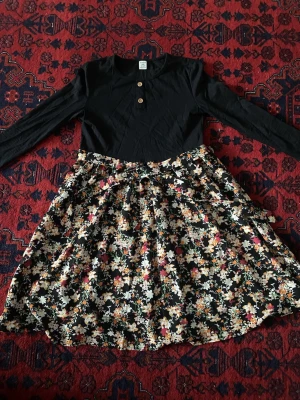 Svart blommig klänning från Shein - Supersöt klänning från Shein med svart överdel, rund hals och två träknappar framtill. Kjoldelen är svart med färgglatt blommönster i vitt, orange och grönt. Långärmad och i mjuk bomull, perfekt för dig som gillar blommiga prints och vill ha en bekväm look.