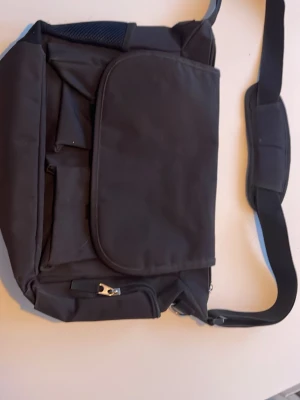 Svart datorväska med axelrem - Säljer en stilren svart datorväska med bred axelrem och vadderad axelkudde för extra komfort. Väskan har flera praktiska fack, dragkedja och ett stort lock framtill. Perfekt för att bära laptop och tillbehör. Enkel och funktionell design.