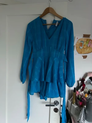 Blå rutig omlottklänning från Amisu - Supersnygg blå kortklänning från Amisu med rutigt mönster, v-ringning och långa ärmar. Klänningen har omlottdetalj i midjan och volang nedtill för en extra flowy look. Perfekt för dig som vill sticka ut med färg och mönster!