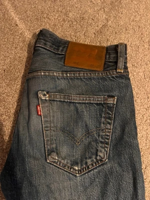 Levi's 501 blå jeans med knappar - Klassiska Levi's 501 jeans i blå tvätt med knappar i gylfen och ikonisk röd Levi's-tag på bakfickan. Jeansen har rak passform och är tillverkade i slitstarkt bomullsjeansmaterial. Perfekt för dig som gillar tidlös denimstil.