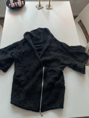 Svart kofta med dragkedja och hålmönster - Cool svart tröja med korta ärmar och hålmönstrad design. Tröjan har en krage och en lång dragkedja framtill. Materialet är stickat och ger en skön känsla. Perfekt för dig som gillar streetstyle och vill sticka ut.