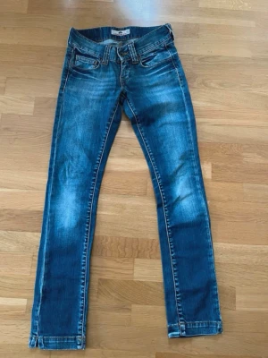 Blå Fornarina jeans i storlek w27 - Säljer ett par blå Fornarina-jeans med klassisk femficksdesign och raka ben. Jeansen har snygga slitningar och kontrastsömmar, samt små slitsar vid bensluten. Modellen är normal i passningen och tillverkad i slitstarkt denimtyg. Perfekt för dig som gillar stilrena jeans med detaljer. Storlek w27 och passar dig med midjemått 68cm. 