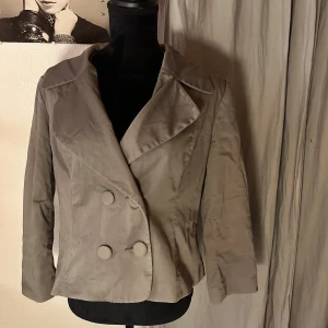 Grå kavaj med stora knappar - Snygg grå kavaj med dubbelknäppning och breda slag. Jackan har långa ärmar och två rader stora, tygklädda knappar framtill. Materialet är slätt och har en lätt glansig finish som ger en cool vibe. Perfekt för dig som gillar stilrena och klassiska plagg.