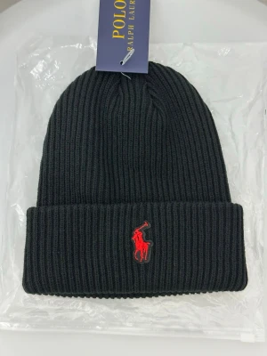 Svart ribbad mössa från Polo Ralph Lauren - Svart ribbstickad mössa från Polo Ralph Lauren med klassisk röd broderad logga framtill. Mössan har uppvikt kant och är tillverkad i mjukt stickat material som håller dig varm och snygg under kyliga dagar.