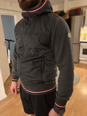 Mörkgrå Moncler bomberjacka med huva - Säljer en mörkgrå bomberjacka från Moncler med huva och dragkedja. Jackan har ribbade muddar och nederkant med röd, vit och marinblå rand. Materialmix med vadderad front i polyester och mjuka ärmar. Klassisk Moncler-logga på ärmen och praktiska fickor med dragkedja.