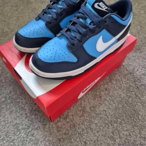 Nike Dunk Low University Blue - Nike Dunk Low GS i färgen University Blue/White. Snygga sneakers med marinblå och ljusblå paneler, vit swoosh och platt sula. Klassisk låg siluett med snörning och perforerad tå. Perfekt för dig som gillar streetwear och vill sticka ut med fräscha färger.
