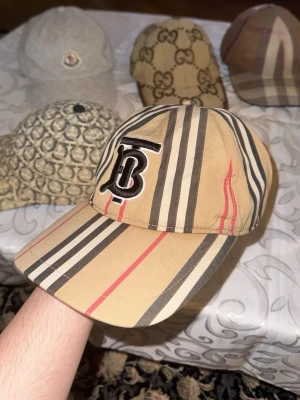 Randig Burberry keps med TB-logga - Säljer en snygg Burberry keps i beige med klassiskt randigt mönster och svarta, vita samt röda detaljer. Framtill sitter en broderad TB-logga. Kepsen har justerbar rem med metallknappar och insida i svart tyg. Perfekt för dig som vill sticka ut.
