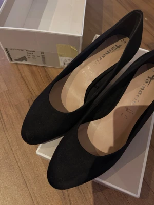 Svarta pumps från Tamaris, storlek 39 - Stilrena svarta pumps från Tamaris i storlek 39  i ett mjukt syntetmaterial med matt finish. Perfekt för dig som gillar klassisk och enkel design med bekväm passform. Nypris 699 säljer för 300kr och priset går att diskutera. Använd en gång. Säljer ett likadant par i storlek 38 också