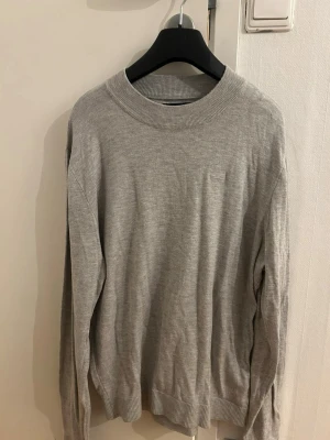 Grå stickad tröja från Hollister - Säljer en stilren grå stickad tröja från Hollister med diskret logga på bröstet. Tröjan har rund hals, ribbade muddar och lång ärm. Perfekt för dig som gillar enkel och klassisk stil.