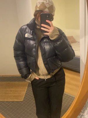 Svart pufferjacka från The North Face - Säljer en svart, croppad pufferjacka från The North Face i storlek M. Jackan har hög krage, dragkedja framtill och snörning i nederkant. Perfekt för kalla dagar och ger en riktigt snygg siluett. Klassisk logga på bröstet och riktigt fluffiga ärmar.