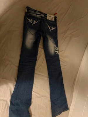 Blå Crazy Age jeans med broderi - Säljer ett par blå jeans från Crazy Age med vita broderade detaljer på både fram- och baksida. Jeansen har en snygg tvätt med slitningar och kontraster, samt raka ben och normal passform. Feta broderier på fickorna och låret ger en unik look.