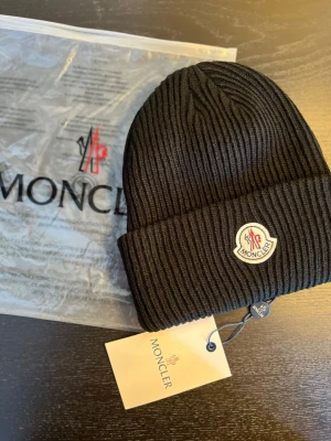 Svart ribbstickad mössa från Moncler - Svart ribbstickad mössa från Moncler med klassisk uppvikt kant och broderad logotyp framtill. Snygg och enkel design som passar till allt, tillverkad i mjukt material som håller dig varm. Perfekt accessoar för kyliga dagar.