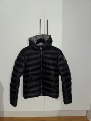 Svart pufferjacka från Colmar - Säljer en svart pufferjacka från Colmar med huva och grått foder. Jackan har en snygg quiltad design, dragkedja framtill och två fickor. Märkeslogga på ärmen ger extra stilpoäng. Perfekt för dig som vill ha en varm och cool jacka till vintern.