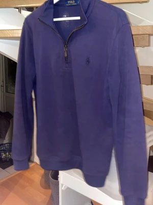 Blå half zip tröja från Polo Ralph Lauren - Säljer en stilren blå långärmad tröja från Polo Ralph Lauren med half zip och liten broderad logga på bröstet. Tröjan har en klassisk krage och är gjord i mjuk bomull, perfekt för dig som gillar enkel och snygg design.