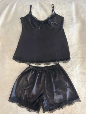 Svart satinsett med spetsdetaljer - Snygg svart sett i satin med tunna axelband och dekorativ rosett framtill. Linnet har spetskant längs urringningen och nederkanten som ger en extra touch. Materialet är glansigt och mjukt, perfekt för dig som gillar en elegant look.