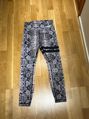 Stronger tränings thights - Stronger träningsbyxor i storlek XS. Materialet är stretchigt & skönt att röra sig i. Byxorna har även en praktisk ficka på midjans baksida. Kan passa vissa mobiler men kan upplevas trång. Perfekt storlek för nycklar till hem & bilen! Upplever att den inte påverkar ens rörelse av att ha saker i fickan. Säljer då de inte längre passar. Sparsamt & sällan använda. Djur och rökfritt hem. Färgerna är en blandning av rosa, lila och svart i ett ormliknande mönster med två svarta streck på vänster lår.