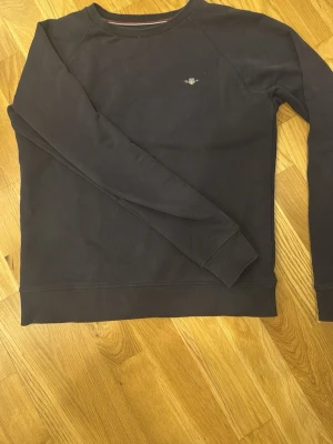 Mörkblå sweatshirt från GANT - Mörkblå långärmad sweatshirt från GANT med diskret logga broderad på bröstet. Tröjan har rund hals med ribbad kant och ribbade muddar vid ärmslut och nederkant. Perfekt för chill dagar och enkel att matcha med jeans eller joggers.