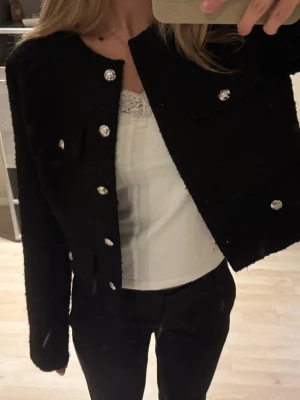 Svart kavajjacka med silver knappar - Snygg svart kavajjacka med silver knappar framtill och på fickorna. Jackan är croppad och har en rak, loose passform med lång ärm. Materialet är mjukt och har en lite ruffad yta som ger en cool vibe. Perfekt att styla med jeans eller kostymbyxor.