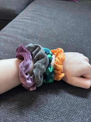 Färgglada scrunchies  - Fyra scrunchies i olika färger: lila, grå, grön och orange. Två är i mjuk sammet och två i glansig satin. Perfekt för att ge din frisyr lite extra färg och stil. Säljes som paket!