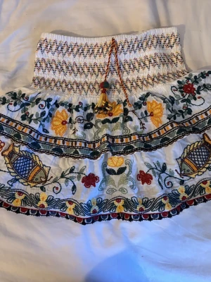 Vit broderad kortkjol från Zara - Supersöt vit kortkjol från Zara med färgglad brodyr i blommönster och fiskar. Kjolen har smock i midjan, dekorativ knytning med tofsar och pärlor samt volangkant. Perfekt för dig som gillar boho-vibbar och unika detaljer.lägg märke till att ena snöret saknar en tofs med pärlor!!