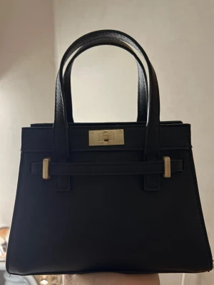 Svart handväska med gulddetaljer - Snygg svart handväska med stilrena gulddetaljer och klassisk fyrkantig form. Väskan har två rejäla handtag och en elegant låsdetalj framtill. Väskan är liten och köpt från asos🥰