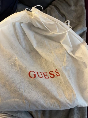 Svart quiltad handväska från Guess - Säljer en svart handväska från Guess med quiltad design och guldiga nitar. Den är i princip oanvänd. Vill få den såld så fort som möjligt, behöver få ut massa ur min garderob så buda gärna! Väskan har både handtag och axelrem, samt guldfärgade detaljer och dragkedja. Insidan är ljus med flera fack och Guess-logga. Perfekt för dig som gillar stilrena accessoarer med lite edge. 