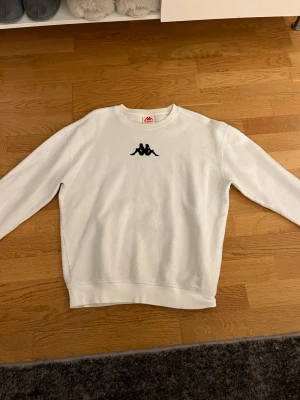 Vit sweatshirt från Kappa - Säljer en stilren vit sweatshirt från Kappa med det klassiska svarta logotrycket på bröstet. Tröjan har rund halsringning, ribbade muddar och är tillverkad i mjukt material som passar perfekt till chill eller streetwear. Inga märken på tröjan förutom det som är med på bild 2, liten miss färgning som inte syns så mycket!