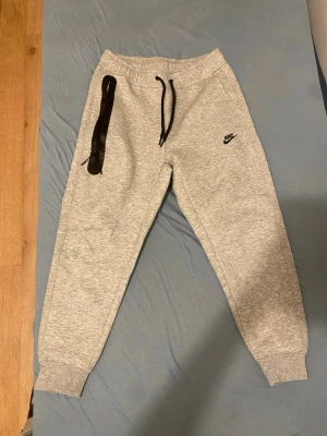 Grå mjukisbyxor från Nike - Säljer ett par grå mjukisbyxor från Nike med svart logga på vänster ben. Byxorna har svarta snören i midjan och en svart dragkedja på höger ficka. Mjukt material och ribbade muddar vid benslut. Perfekta för chill eller träning.