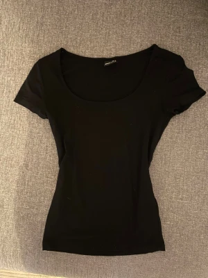 Svart topp från Gina Tricot XS - En enkel svart topp från Gina Tricot i storlek XS. Modellen har rundad halsringning och korta ärmar, med en slim passform som sitter snyggt mot kroppen. Perfekt att matcha med jeans eller kjol för en clean look.