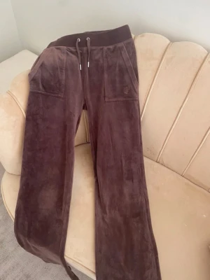 Bruna mjukisbyxor Juicy Couture XS - Bruna mjukisbyxor från Juicy Couture i storlek XS. Byxorna har bred resår i midjan med snörning, stora fickor fram och bak med tryckknappar. Materialet är mjukt och glansigt, typiskt för sammet. Perfekt för chill och comfy vibes. Dom är i ett jättebra skick, har inte använt på några år. Dom är lite slitna längst ner. Pris kan sänkas vid snabb affär 
